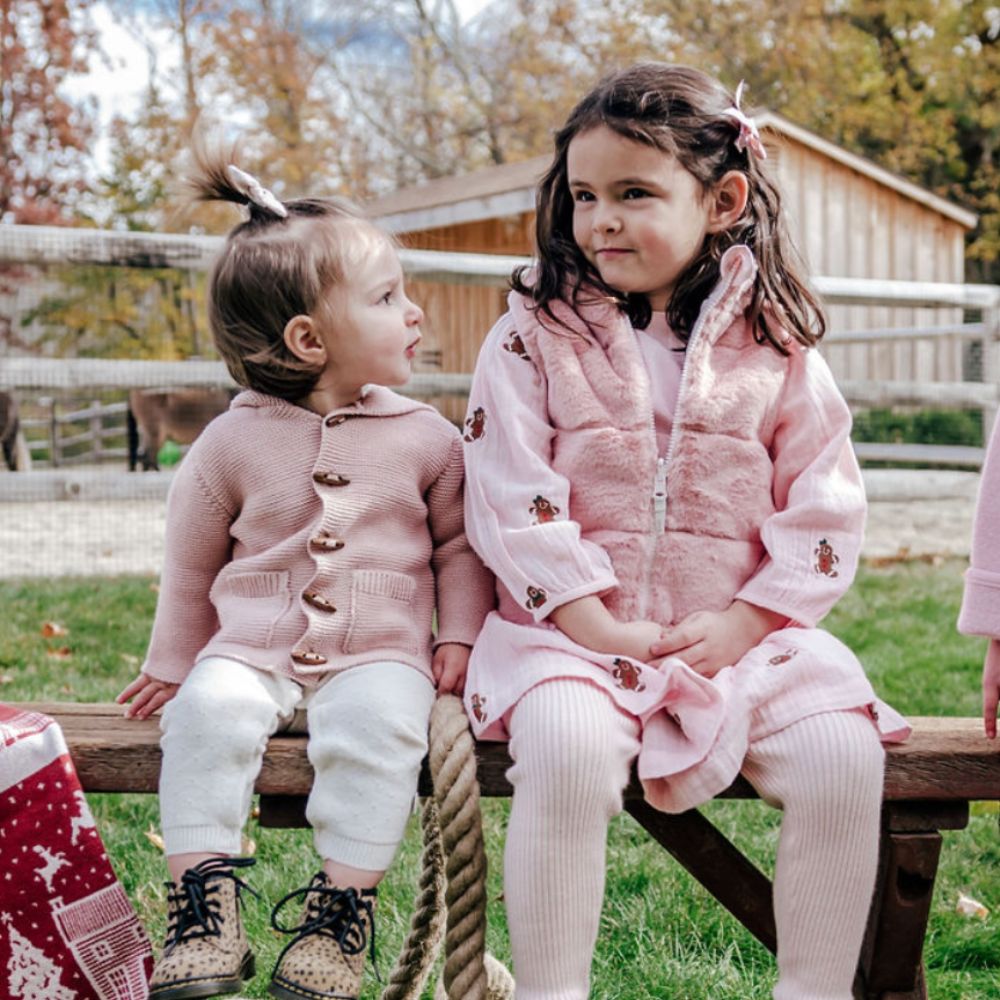 two girls on bench with pink sweaters and a fur vest